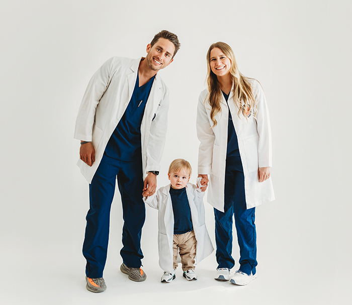 Doctors Kyle and Kasey Kringle holding hands with their toddler all in white lab coats